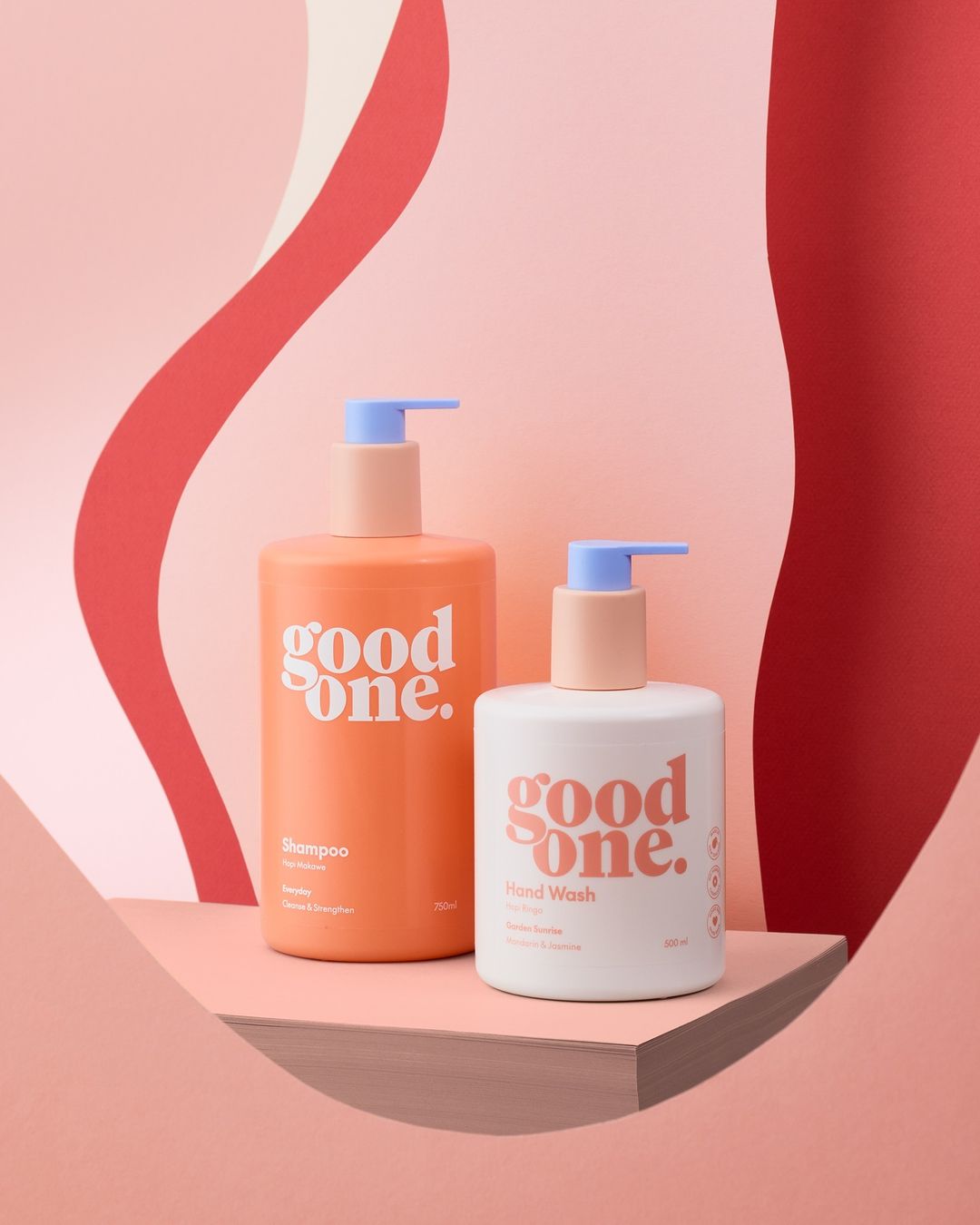 Two pastel-colored bottles of Good One brand shampoo and hand wash are staged in a studio with a peach-toned platform and background.