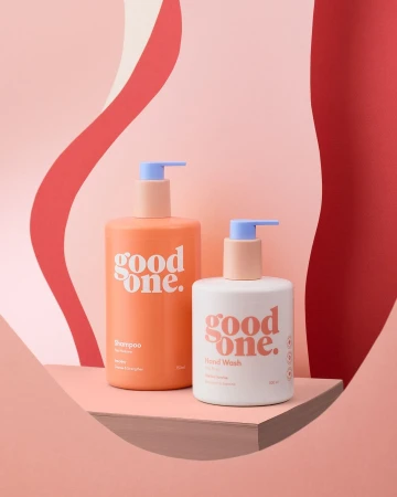 Two pastel-colored bottles of Good One brand shampoo and hand wash are staged in a studio with a peach-toned platform and background.