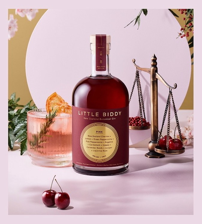A bottle of Little Biddy Gin with a glass of a mixed drink, garnished with a slice of orange, and a set of scales with cherries.