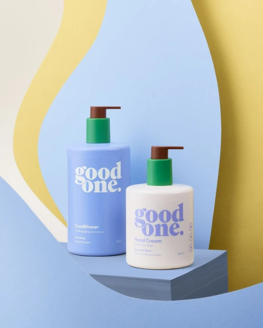 Two 'Good One' brand personal care bottles on a blue platform, against a background of yellow and light blue curved panels.