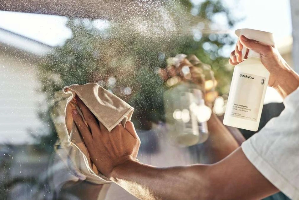 Person cleaning a window with a cloth and cleaning spray. White spray bottle and cleaning cloth used for window cleaning, indoor environment.