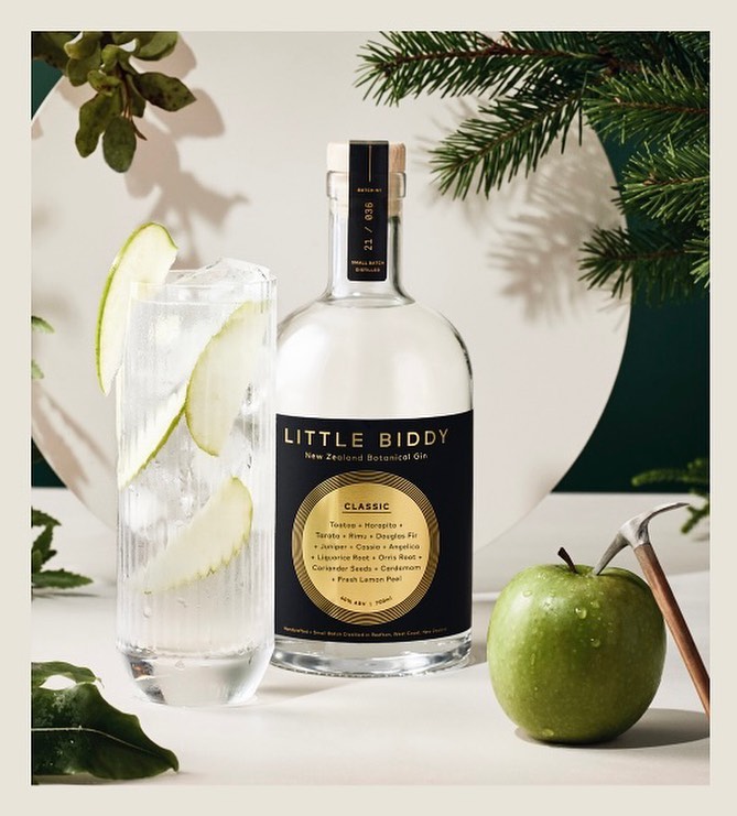 Still life shot of Little Biddy Gin bottle, apple and a gin and tonic drink, set against a white backdrop and green foliage.