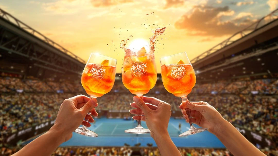 Three Aperol Spritz drinks are held in front of a stadium crowd, with a bright sun and orange sky visible in the background.