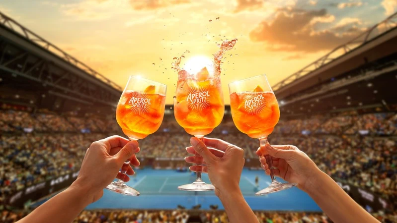Three Aperol Spritz drinks are held in front of a stadium crowd, with a bright sun and orange sky visible in the background.