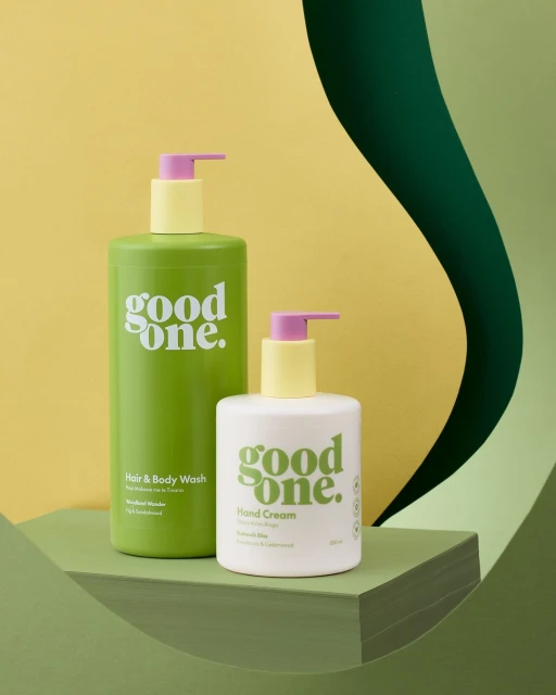 Still life shot of Good One brand hair and body wash and hand cream bottles with pastel pumps against a green and yellow background.