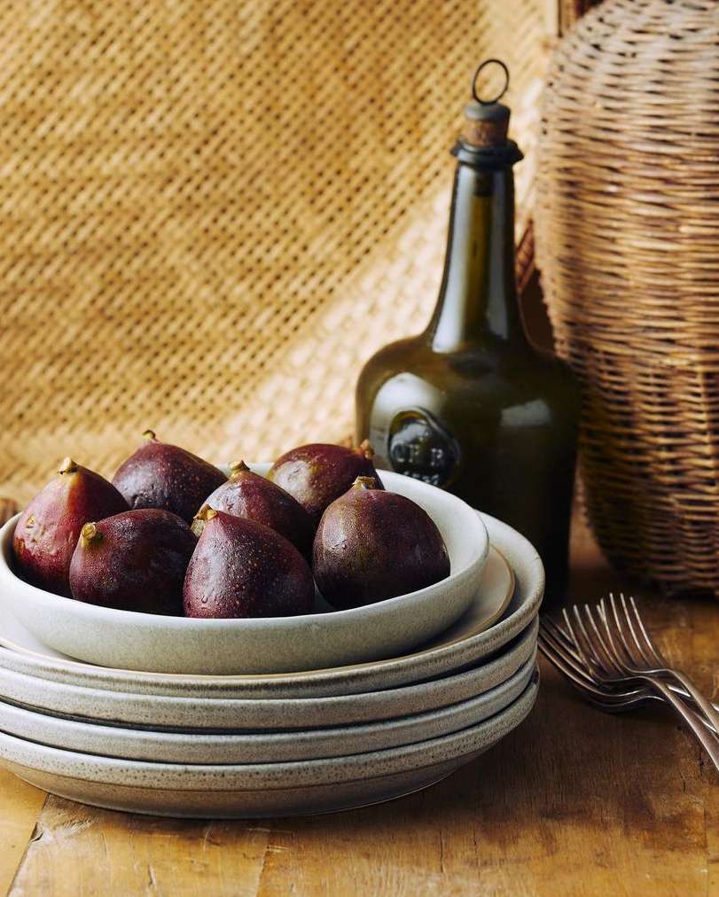 Still life featuring figs in a bowl on stacked plates, alongside a green glass bottle and silverware, set against a woven background.