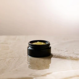 Black cosmetic jar with green cream resting on a reflective wet textured stone surface