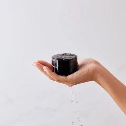 Water droplets falling onto a dark cosmetic jar resting in an open palm against a white background.
