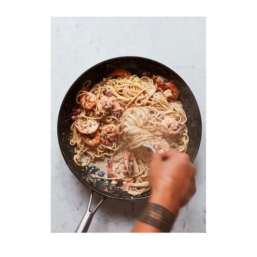 Overhead shot of shrimp pasta being prepared in a black pan on a marble surface, featuring plump shrimp and creamy sauce.