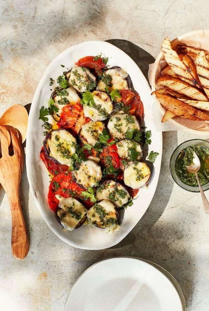 Overhead view of caprese salad with grilled eggplant, sliced tomatoes, mozzarella, pesto, french fries and white plates.