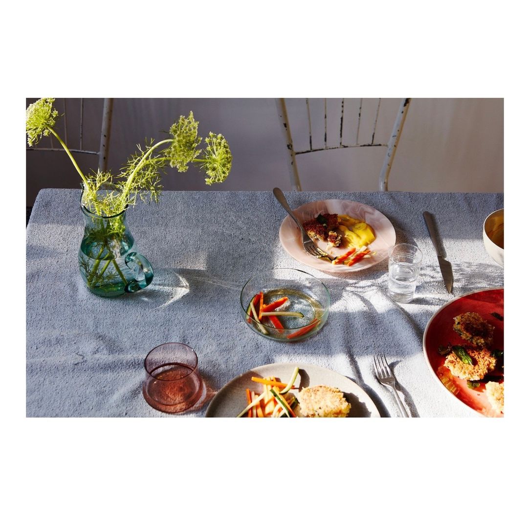 Still life photograph of a table set with plates of food, glasses, utensils, and a vase of flowers under natural light.