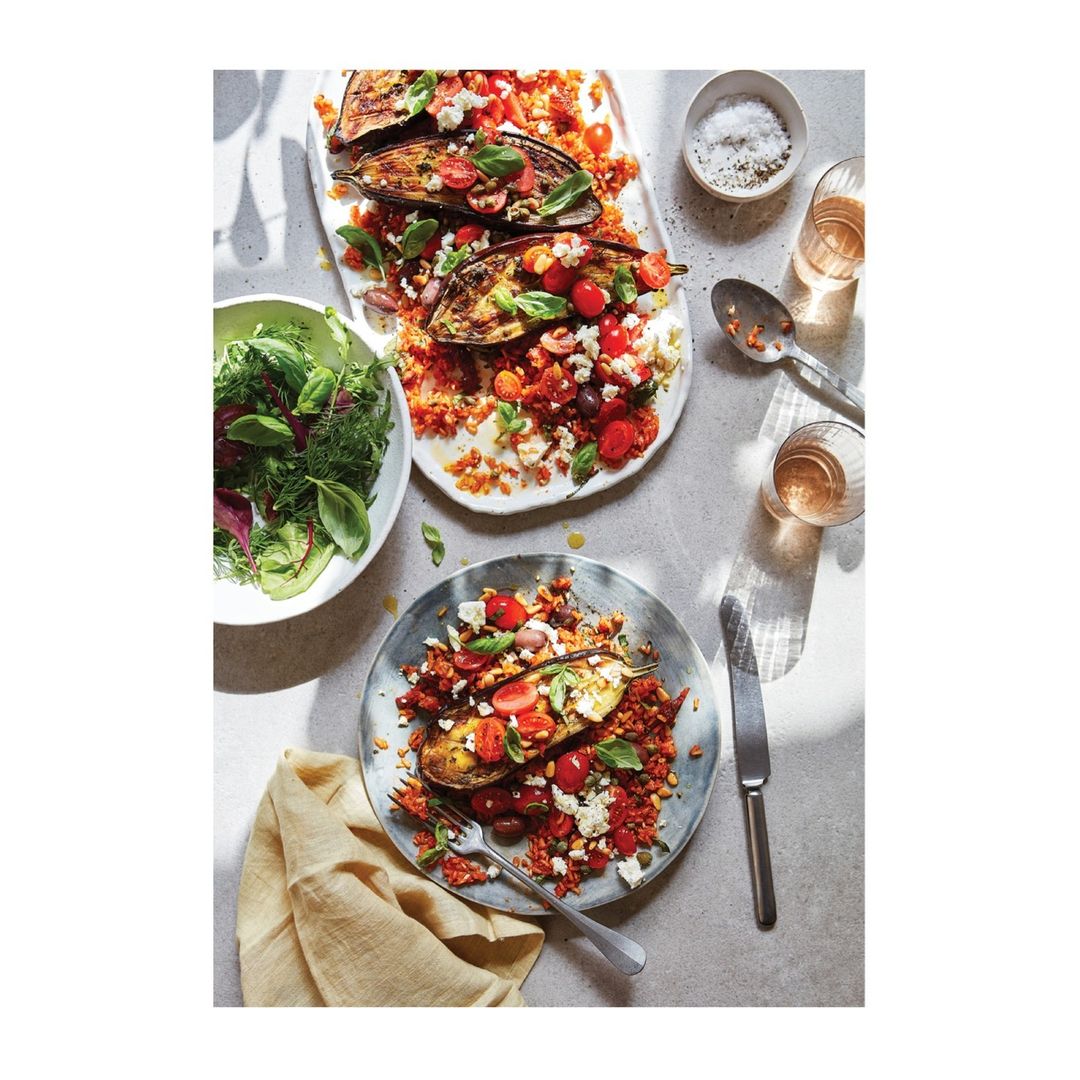 Overhead shot of plates and bowls filled with eggplant, tomatoes, fresh basil, and couscous, with drinks and silverware on a gray surface.