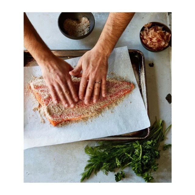 Overhead shot of salmon fillet being coated in breadcrumbs by a person, with cooking tray, parchment paper, herbs, and spices.