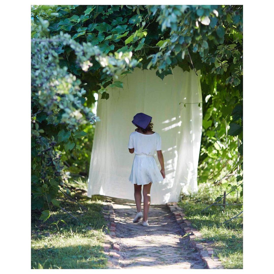 Woman in white dress and blue headscarf walking on brick path in a garden setting with green foliage and a white sheet hanging.