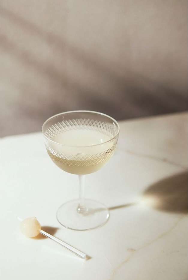 Elegant cocktail in a coupe glass with a small spherical garnish on a clear stick, presented on a white marble surface with soft lighting.