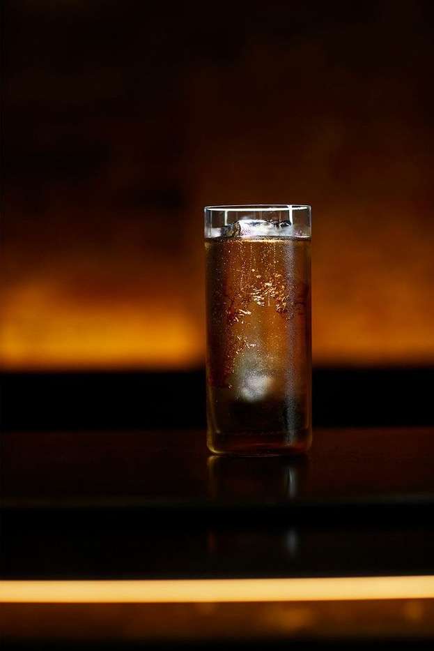 A tall clear glass filled with a brown beverage and ice sits on a glossy black surface, against a blurred brown background.