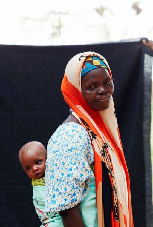 An African woman carries a baby on her back, wrapped in cloth. The woman wears a patterned headscarf and a floral print shirt.