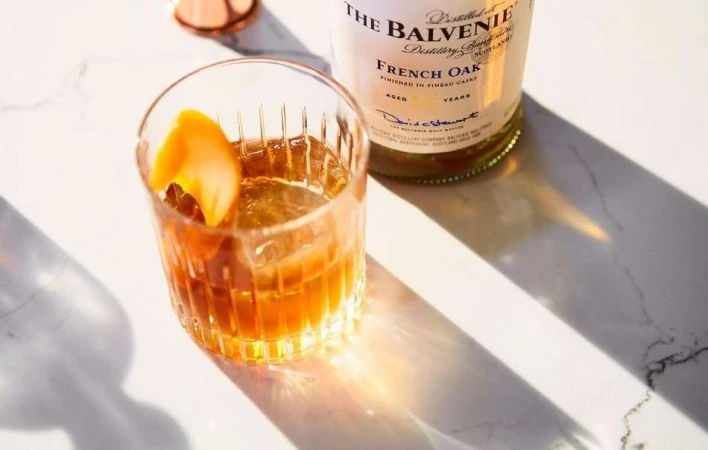 Cocktail in a glass with an orange twist and a bottle of whiskey on a marble table with sunlight and shadow.