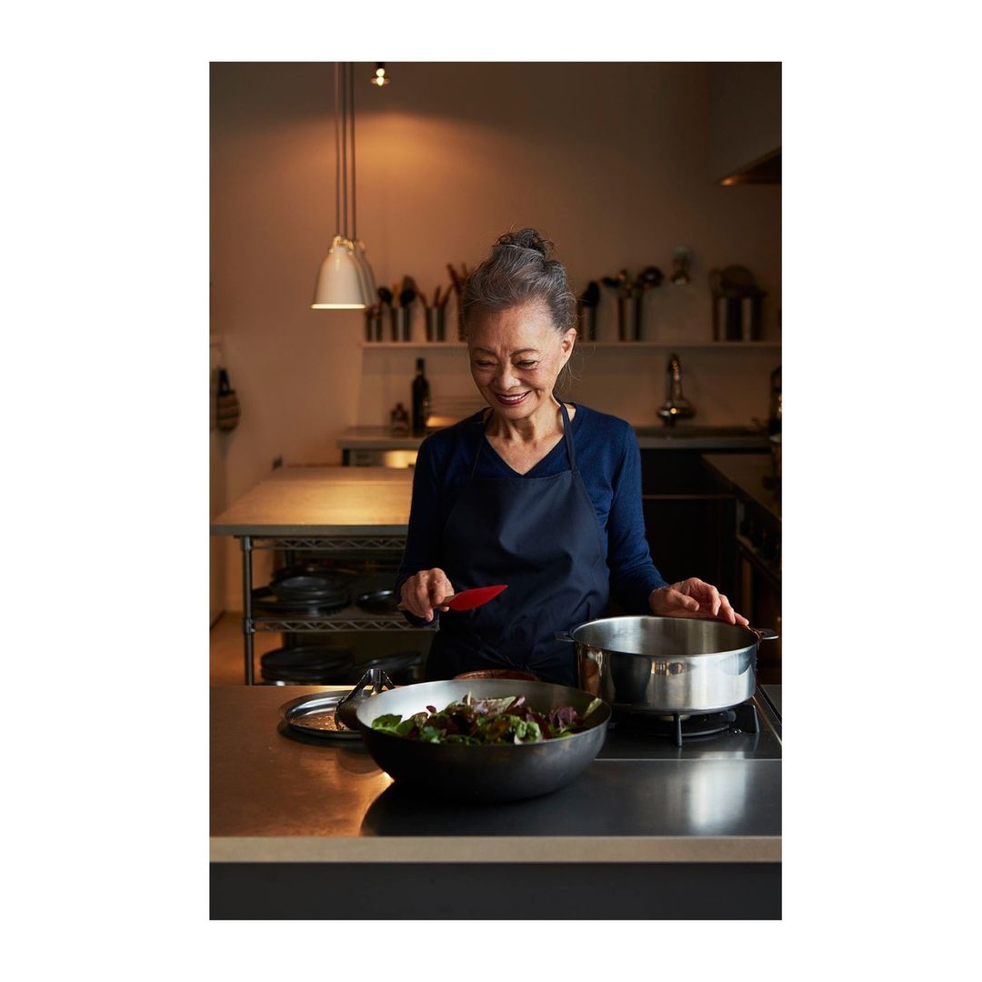 Older adult woman cooking in a kitchen. She is wearing an apron and stirring a bowl of salad next to a pot on a stainless steel counter.