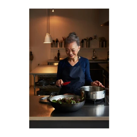 Older adult woman cooking in a kitchen. She is wearing an apron and stirring a bowl of salad next to a pot on a stainless steel counter.