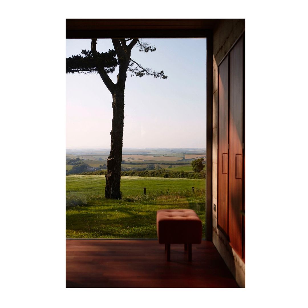 Interior shot of a room with a wooden floor and a view of a green field and tree through a large window. Minimalist bench in the room.