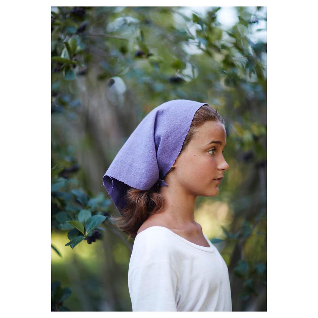 Profile shot of a teenage girl outdoors wearing a lavender headscarf and white t-shirt, with a leafy green background.