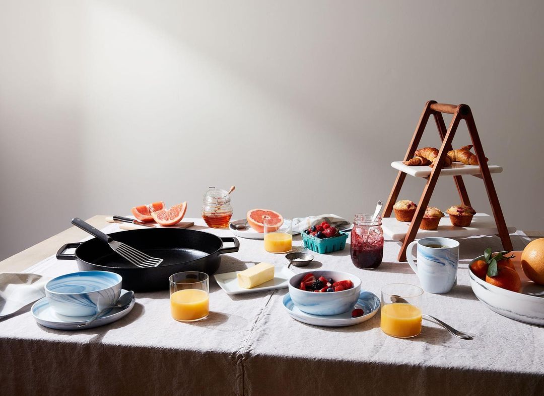 Breakfast table featuring a cast iron pan, assorted fruits, pastries, juice, and marmalade, all set on a linen tablecloth.