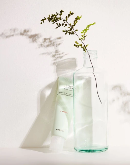 Still life shot of vegan sunscreen tube propped on a white surface next to a clear glass bottle with greenery inside.