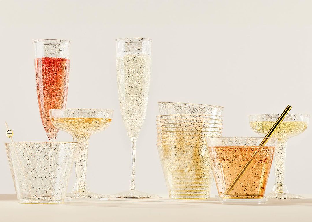 Several clear plastic glasses in various shapes filled with colorful sparkling beverages are arranged on a light surface.