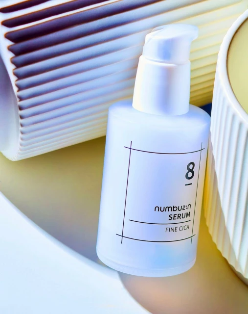 Cosmetic bottle of Numbuzin serum surrounded by decorative objects in cream and white hues on a light surface, product shot.