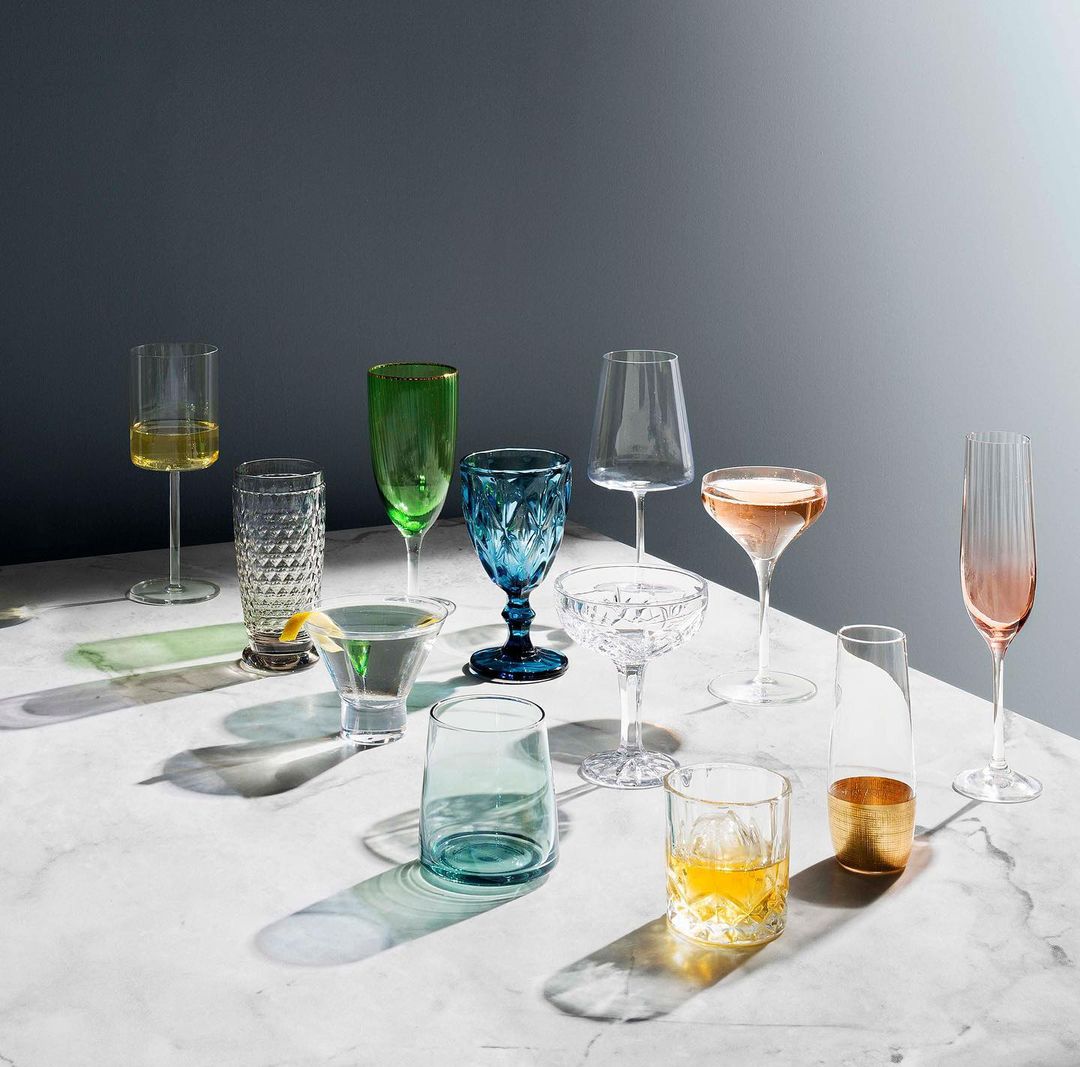 Elegant arrangement of assorted colorful glassware on a marble surface, showcasing various shapes and drink types against a gray background.