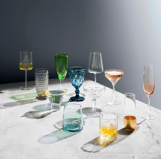 Elegant arrangement of assorted colorful glassware on a marble surface, showcasing various shapes and drink types against a gray background.