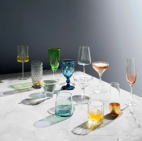 Elegant arrangement of assorted colorful glassware on a marble surface, showcasing various shapes and drink types against a gray background.