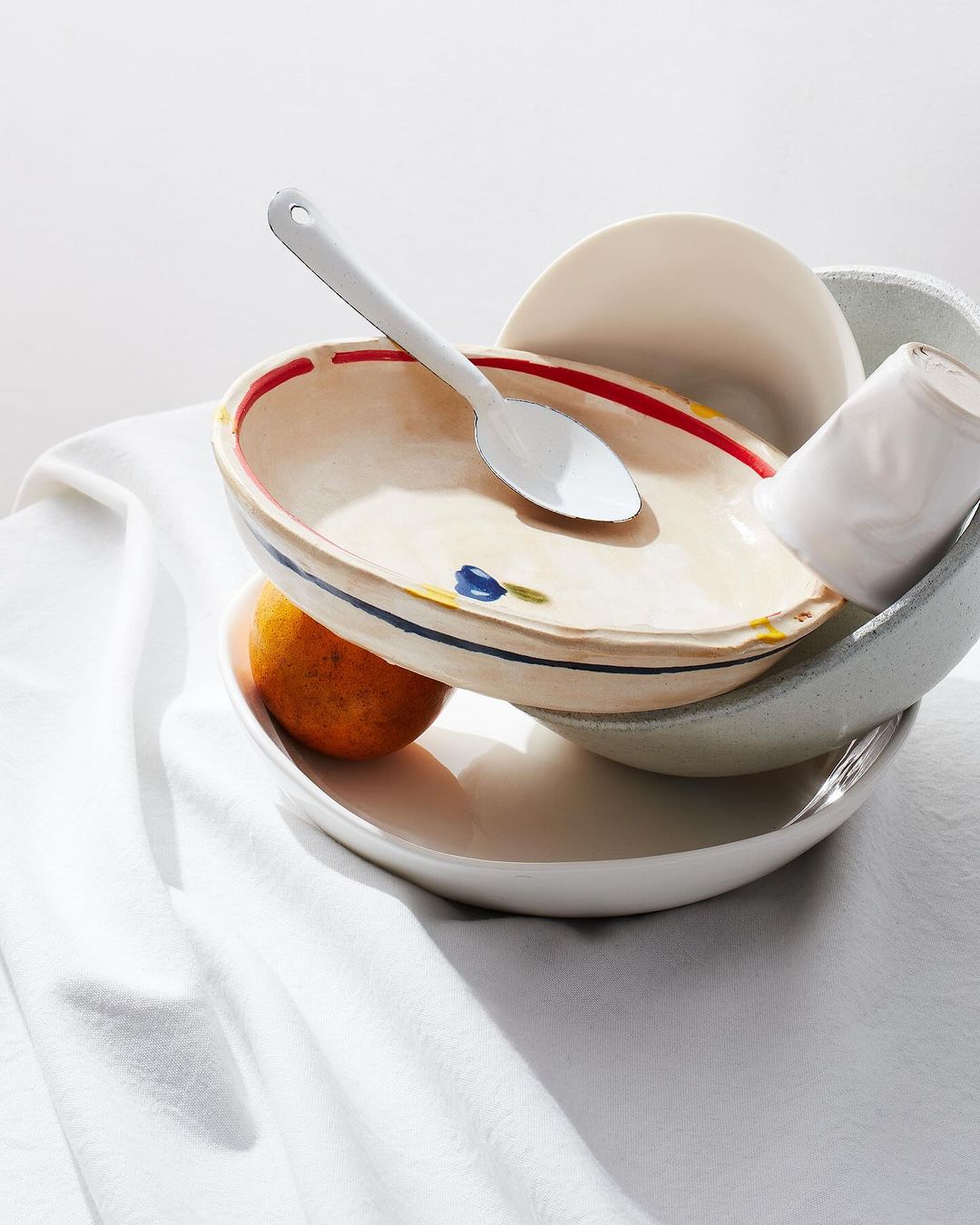 Still life photo of ceramic bowls stacked on a white cloth with a spoon and tangerine, in bright light for food and beverage content.
