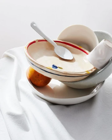 Still life photo of ceramic bowls stacked on a white cloth with a spoon and tangerine, in bright light for food and beverage content.