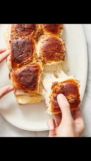Close-up shot of hands pulling apart ham and cheese sliders with mustard seeds on a white plate, showcasing gooey cheese.
