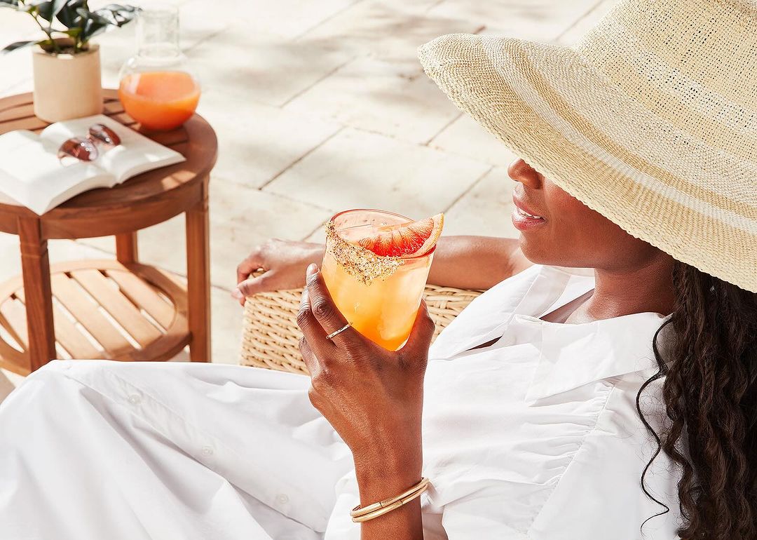 Woman in sunhat holds citrus cocktail outside on patio, sitting next to a wooden table with a drink pitcher and open book.