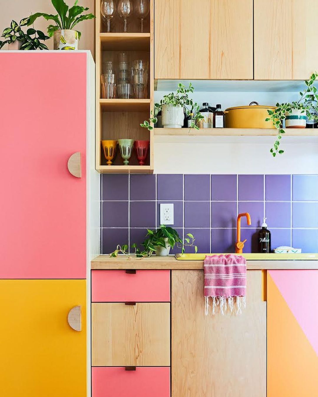 Colorful kitchen interior with pink and yellow cabinets, purple backsplash, wooden countertops, plants, glassware, and copper faucet.
