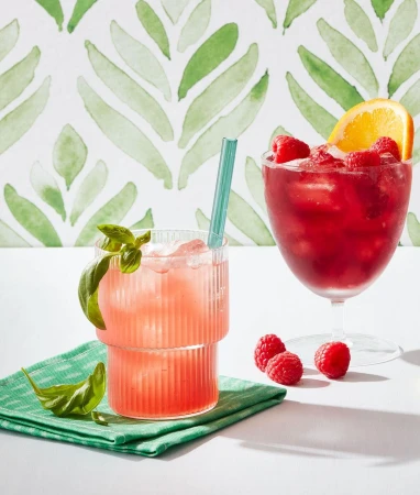 Two cocktails in clear glasses with raspberry and orange garnish. One cocktail has basil leaves, sitting on a green cloth napkin.