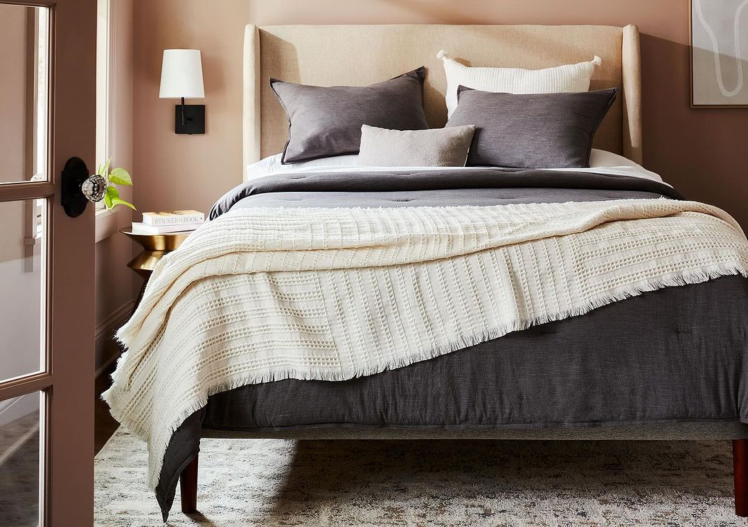 Bedroom features a gray upholstered bed, neutral walls, a throw blanket, pillows, wall sconce, and a side table with decor.