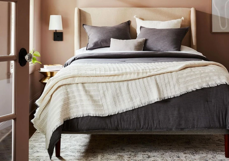 Bedroom features a gray upholstered bed, neutral walls, a throw blanket, pillows, wall sconce, and a side table with decor.