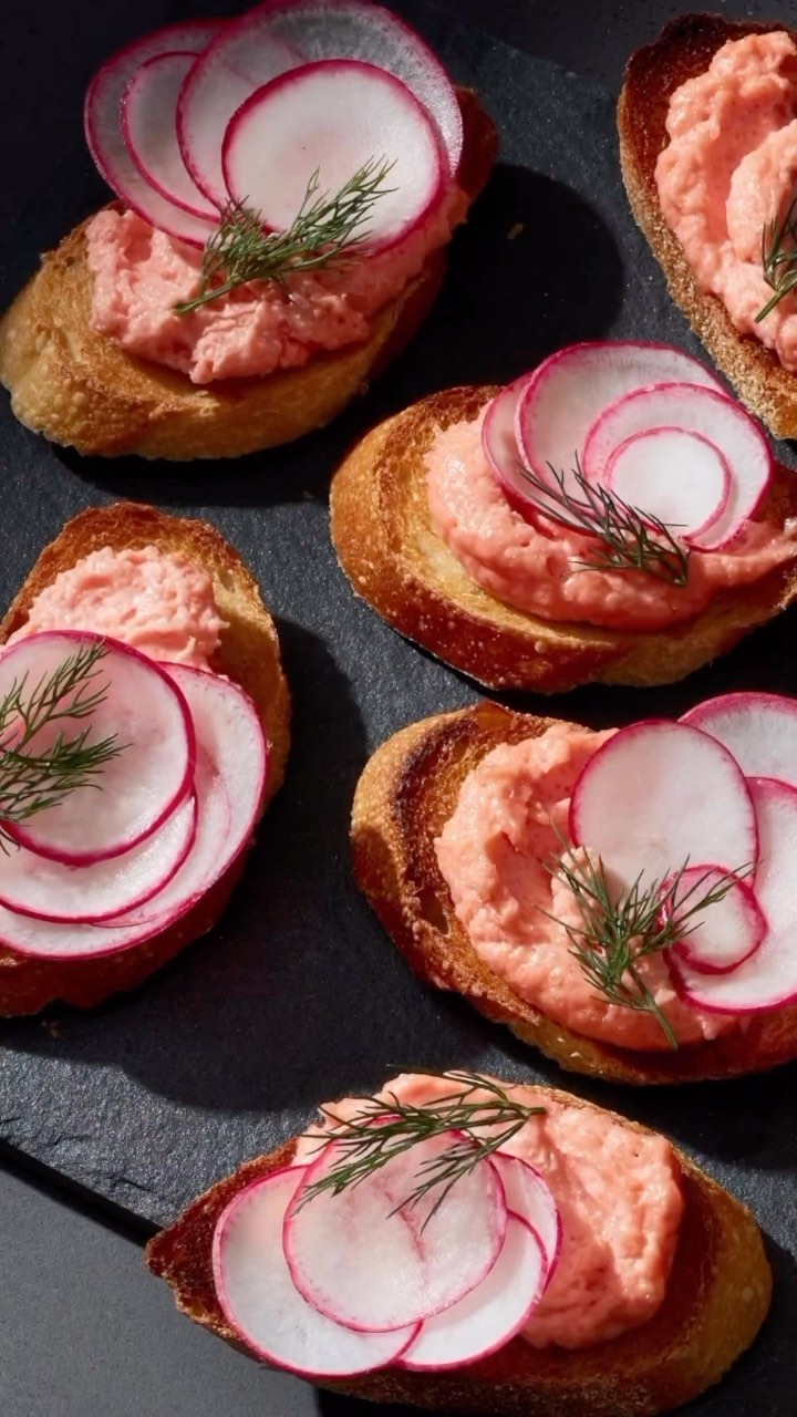 Overhead shot of several crostini appetizers with pink topping and radish slices on a dark surface, food photography. 
