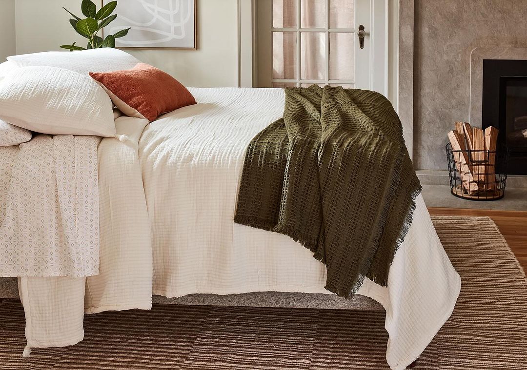 Cozy bedroom interior featuring a made bed with white linen, textured throw, decorative pillow, and a wire basket with logs.