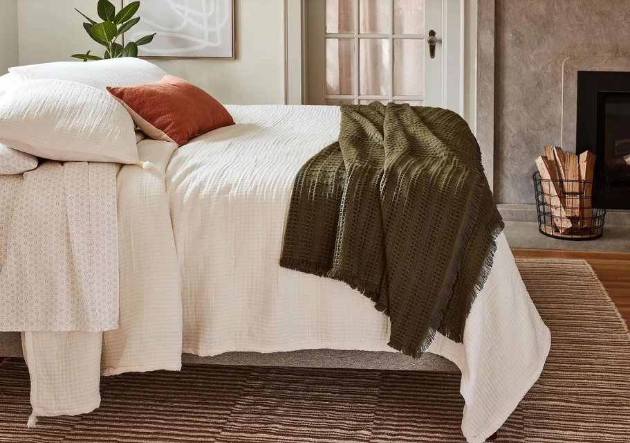 Cozy bedroom interior featuring a made bed with white linen, textured throw, decorative pillow, and a wire basket with logs.