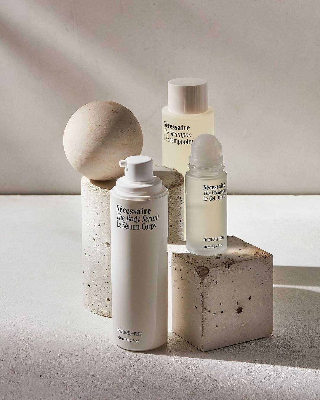 Still life shot of NÉCESSAIRE brand body serum, shampoo, and gel deodorant on concrete pedestals against a neutral background with shadow play.