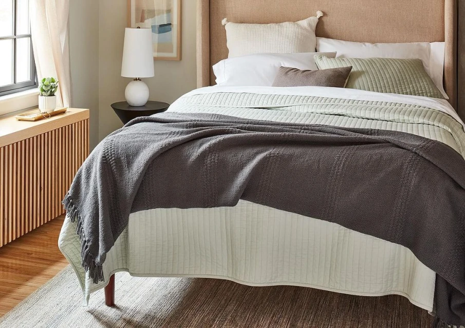 Bedroom interior featuring a neatly made bed with a textured grey throw, white linens, and decorative pillows in a neutral color scheme.