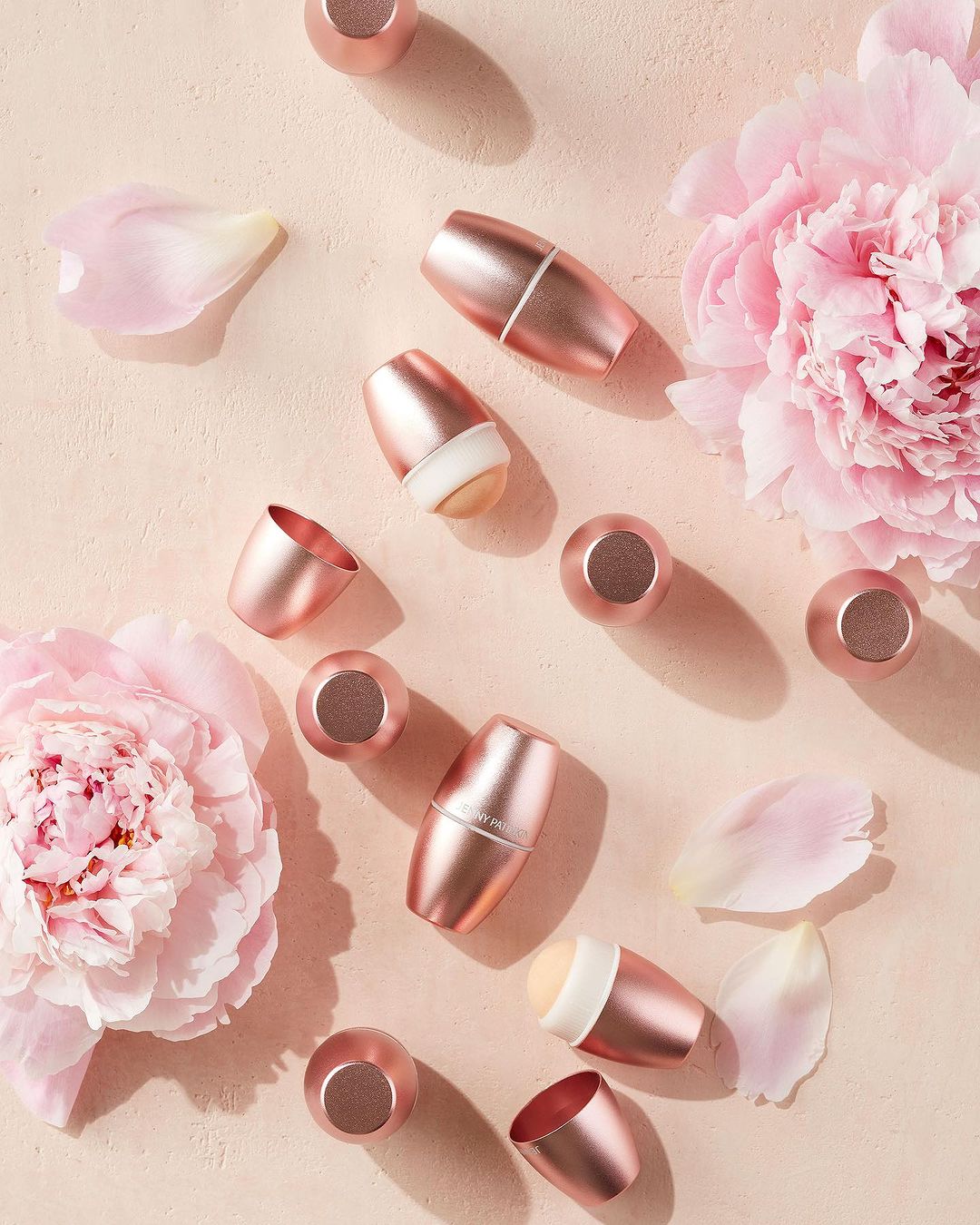 Overhead shot of pink makeup applicators with detachable heads, surrounded by peony blossoms and petals on a light beige surface.