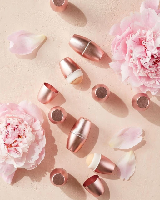 Overhead shot of pink makeup applicators with detachable heads, surrounded by peony blossoms and petals on a light beige surface.