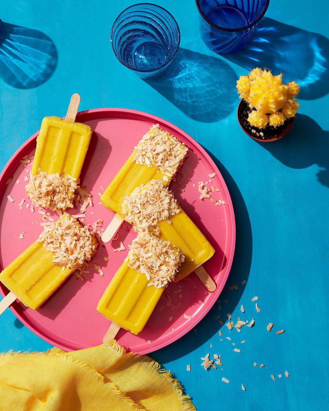 Overhead shot of several yellow popsicles topped with shredded coconut on a pink plate, set against a vibrant blue background.