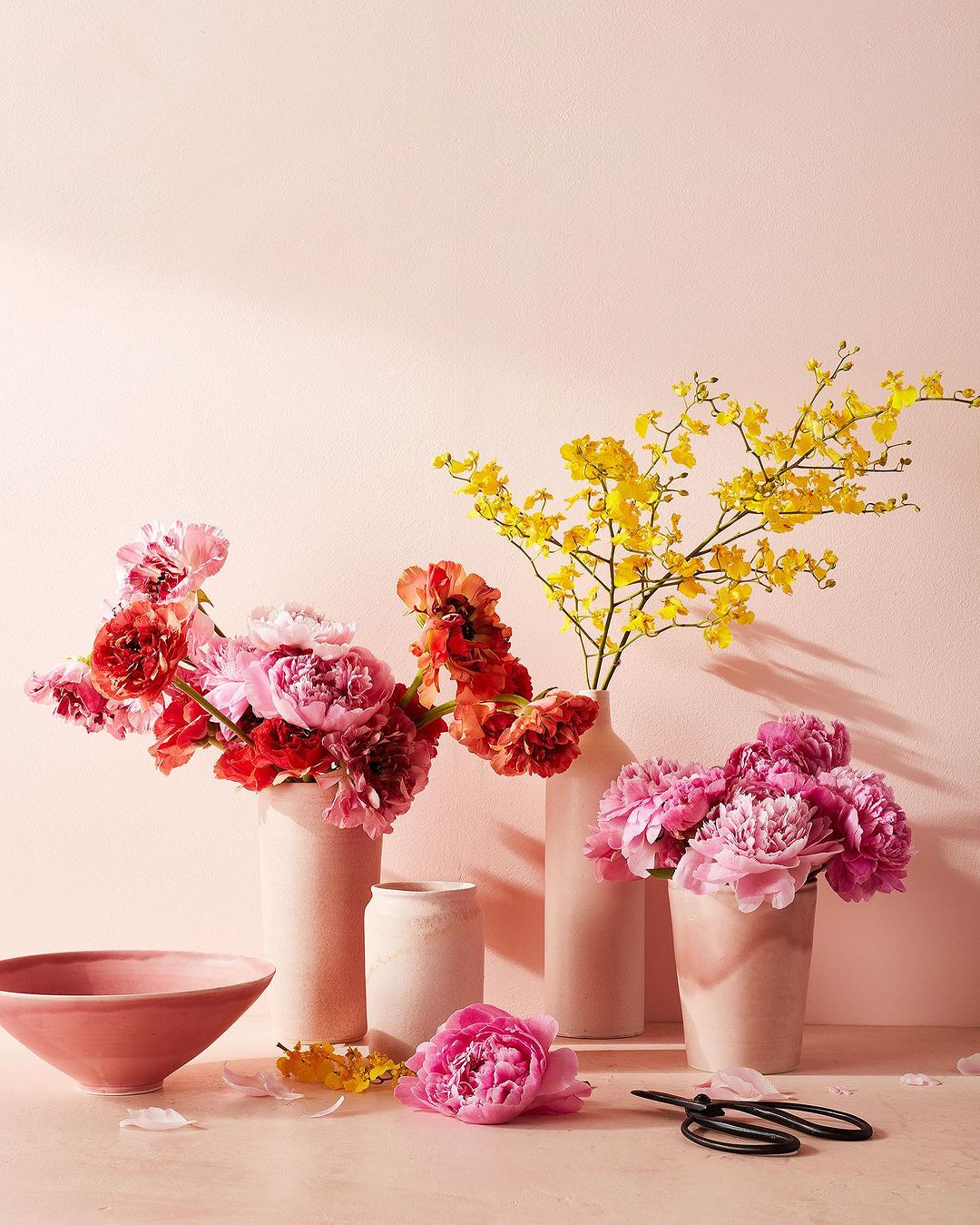Still life of pastel-colored peonies and yellow oncidium orchids arranged in ceramic vases, bowl, and a pair of scissors on table.
